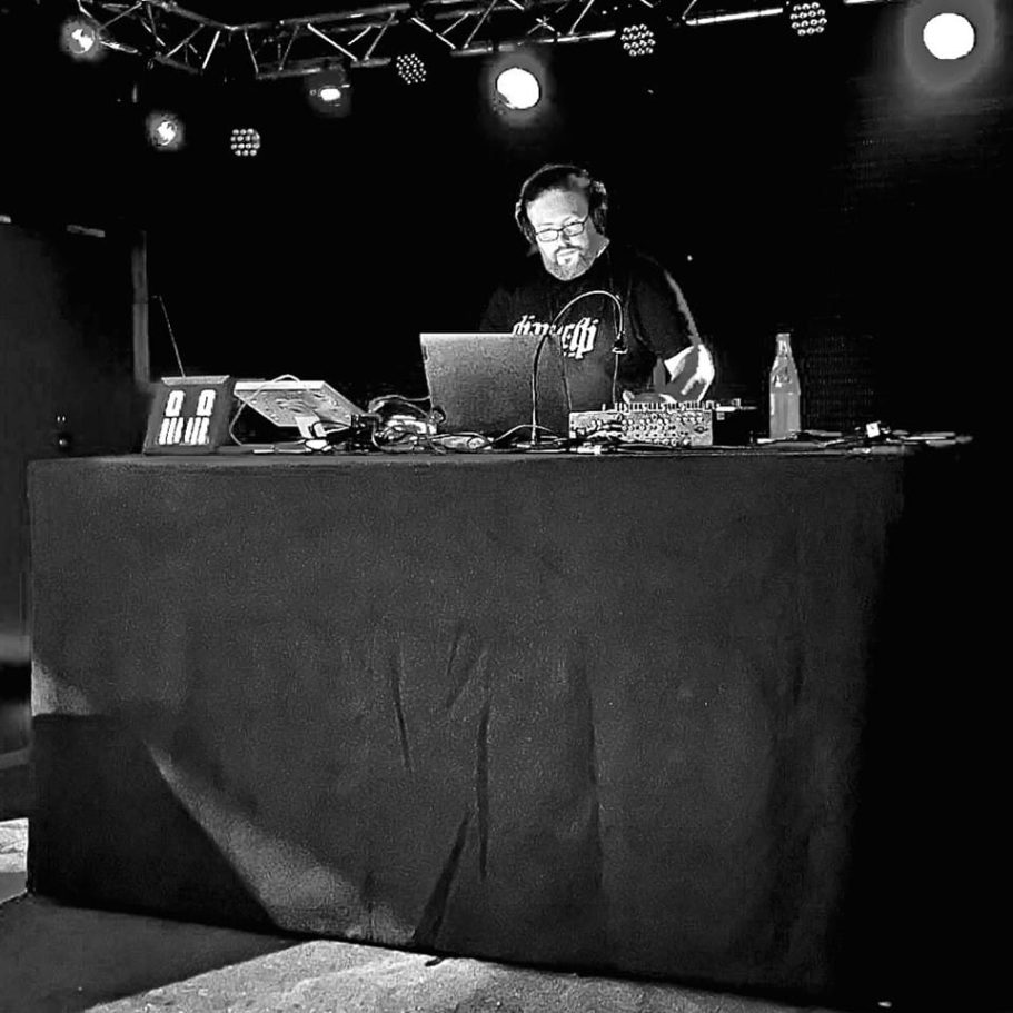 DJ steht an einem Tisch, umgeben von Technik und Beleuchtung in einem dunklen Raum.