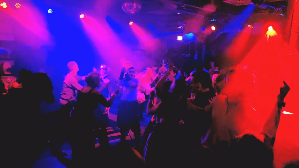 Tanzende Menschen in einem lebhaften, bunten Club mit Lichtern und Nebel.